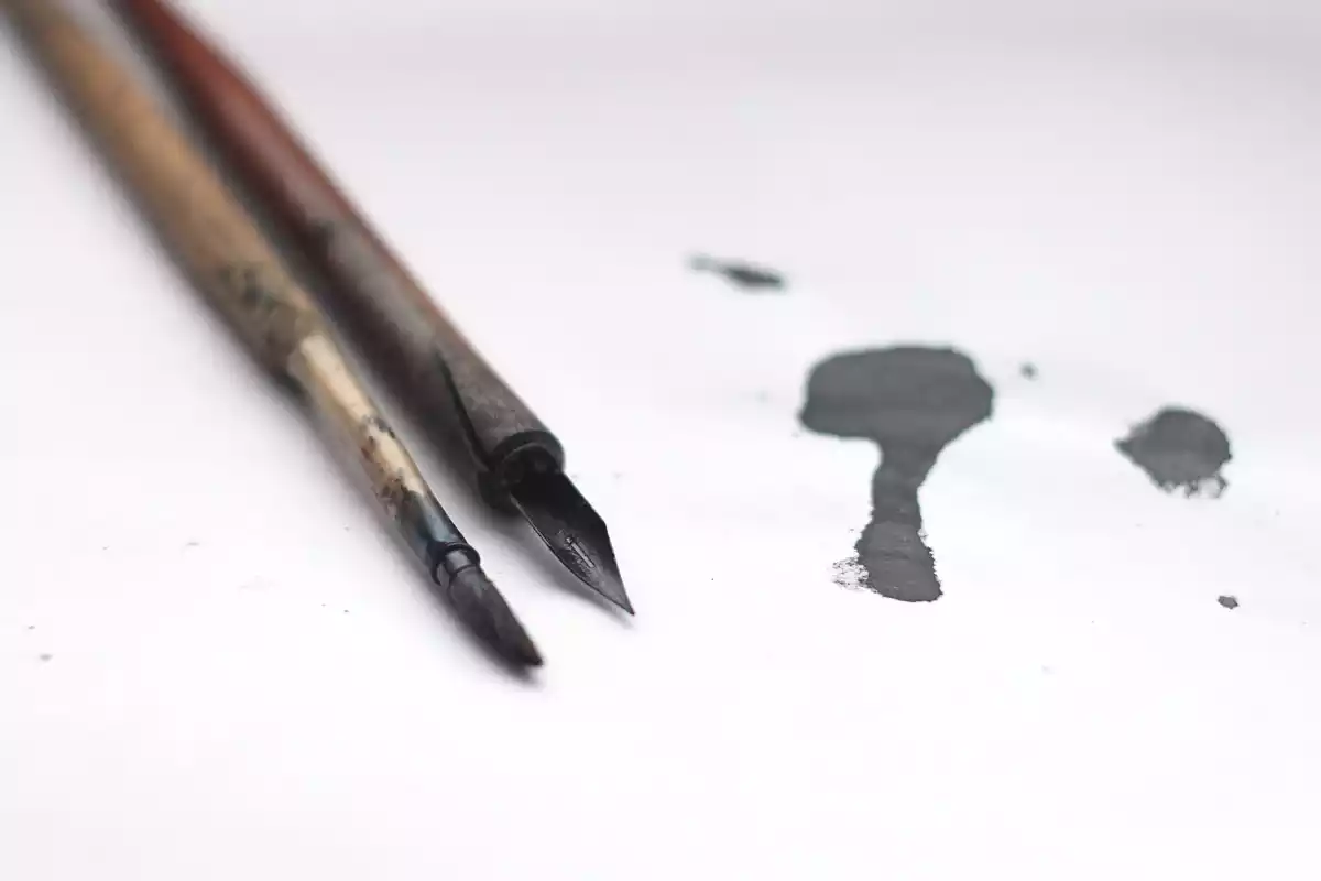 Un bolígrafo y un pincel al lado de una mancha de tinta sobre un papel blanco