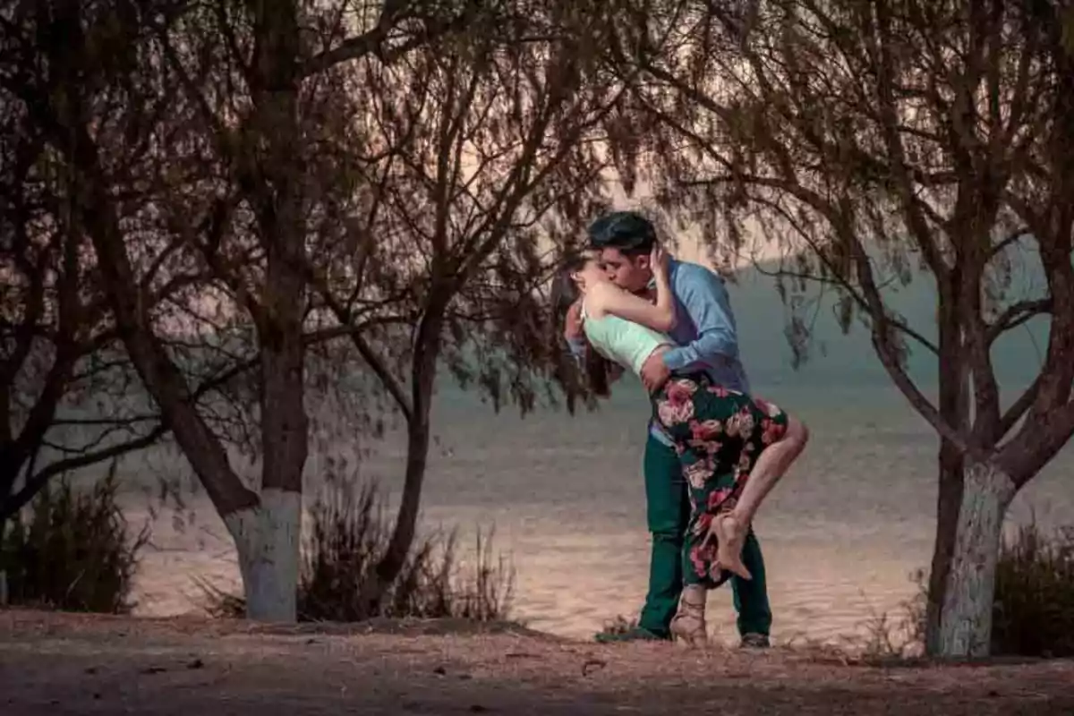 Una pareja besándose apasionadamente en un entorno natural con árboles y un lago al fondo.