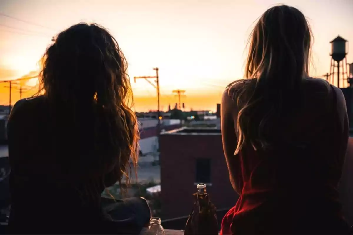 Dos personas sentadas de espaldas observando el atardecer en una ciudad con botellas a su lado.