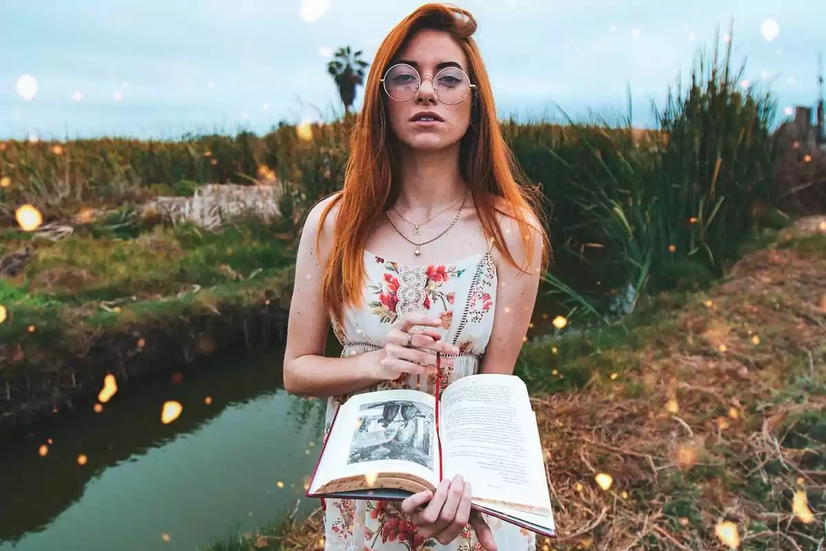 Mujer con cabello largo y pelirrojo, usando gafas y un vestido floral, sostiene un libro abierto mientras está de pie en un entorno natural con vegetación y un pequeño canal de agua.