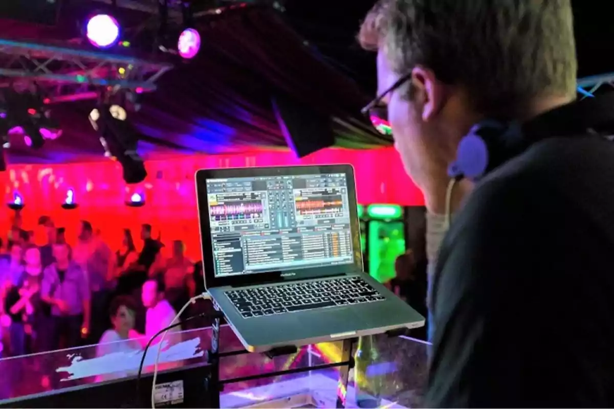 Un DJ mezclando música en una fiesta con luces de colores y una multitud en el fondo.