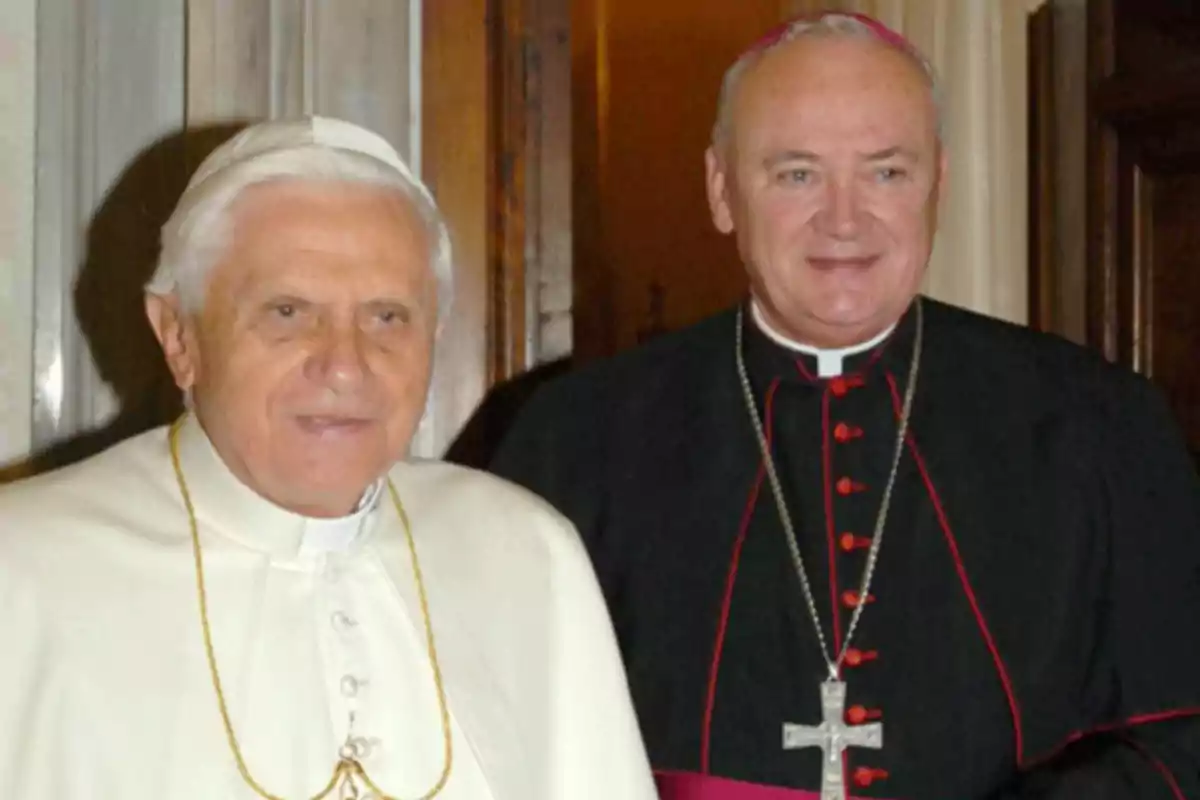 Dos clérigos católicos posan juntos, uno vestido de blanco y el otro de negro con detalles rojos.