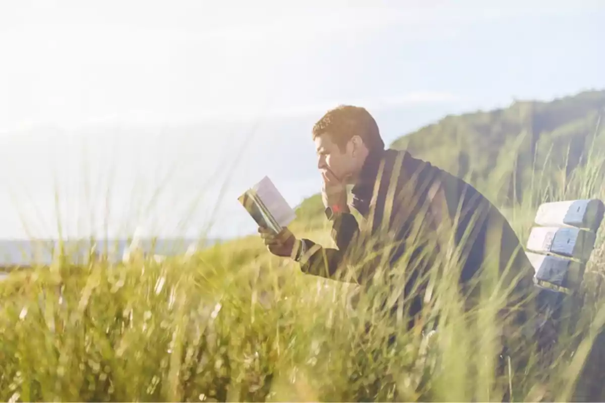 Hombre sentado en un banco leyendo un libro en un campo con hierba alta y colinas al fondo.