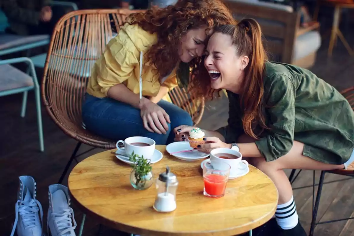 Dos mujeres riendo y disfrutando de una merienda en una cafetería, con tazas de café, un cupcake y un vaso de jugo sobre la mesa.