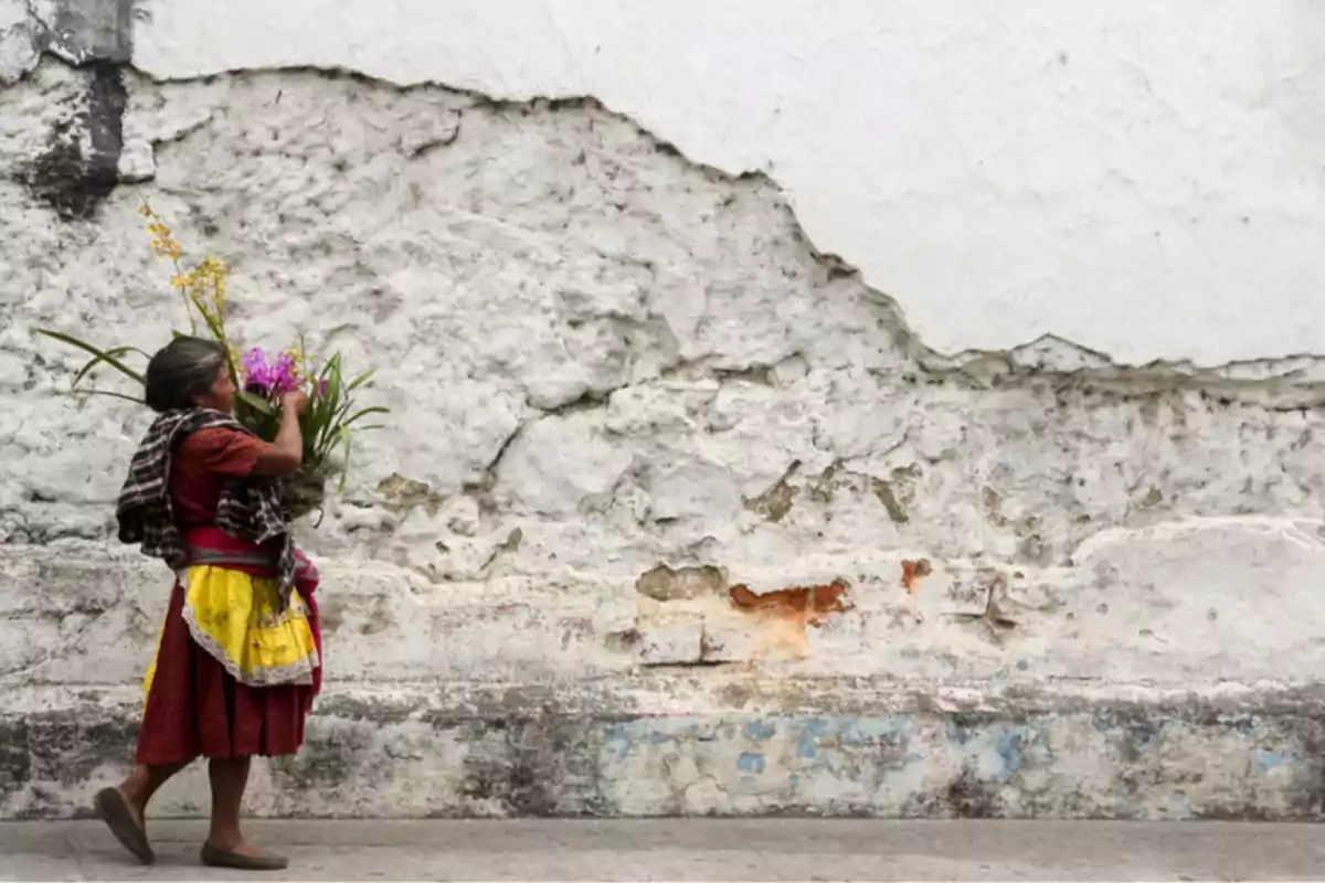 Una mujer con ropa tradicional camina junto a una pared desgastada mientras lleva flores en sus manos.