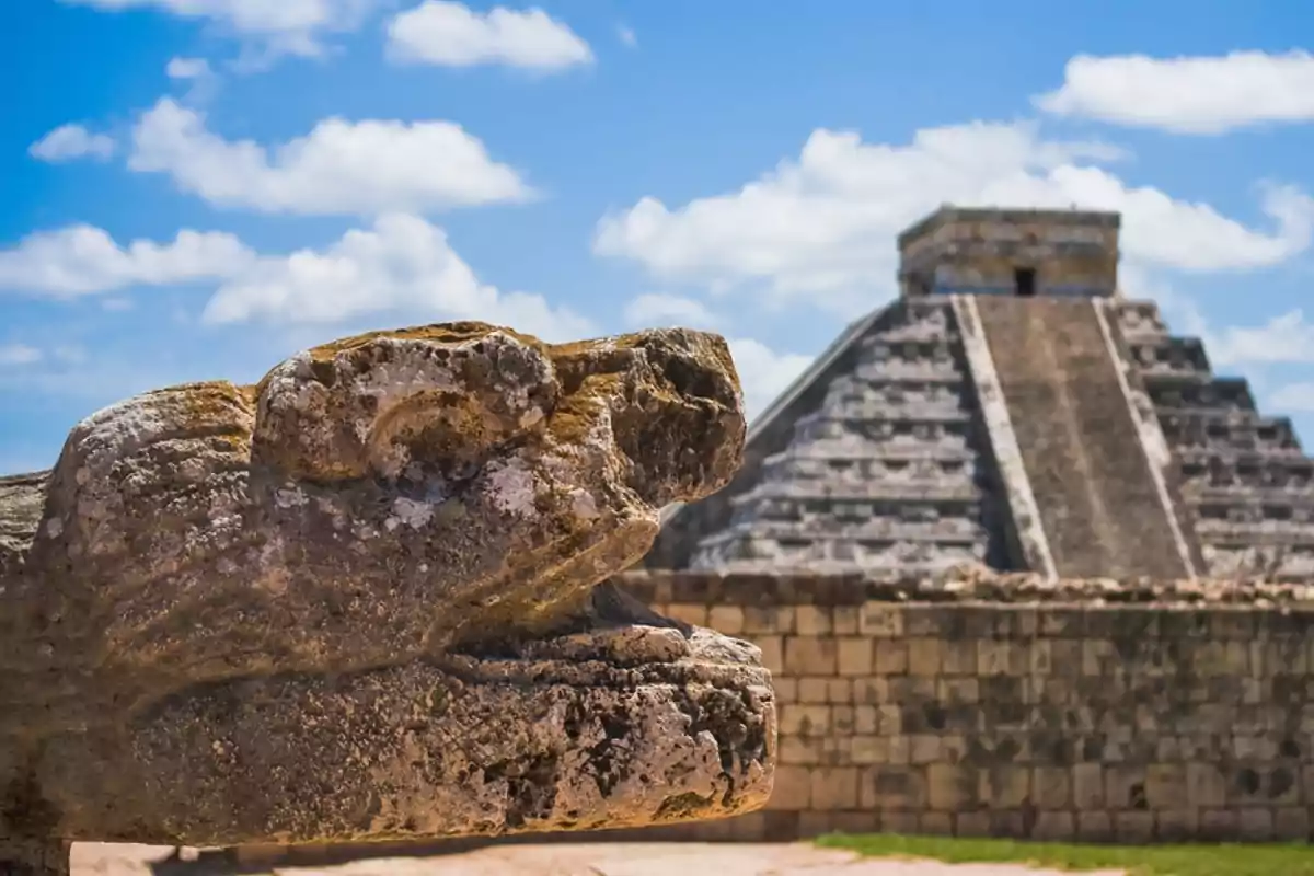 Escultura de piedra en forma de serpiente con la pirámide de Chichén Itzá al fondo bajo un cielo azul con nubes.