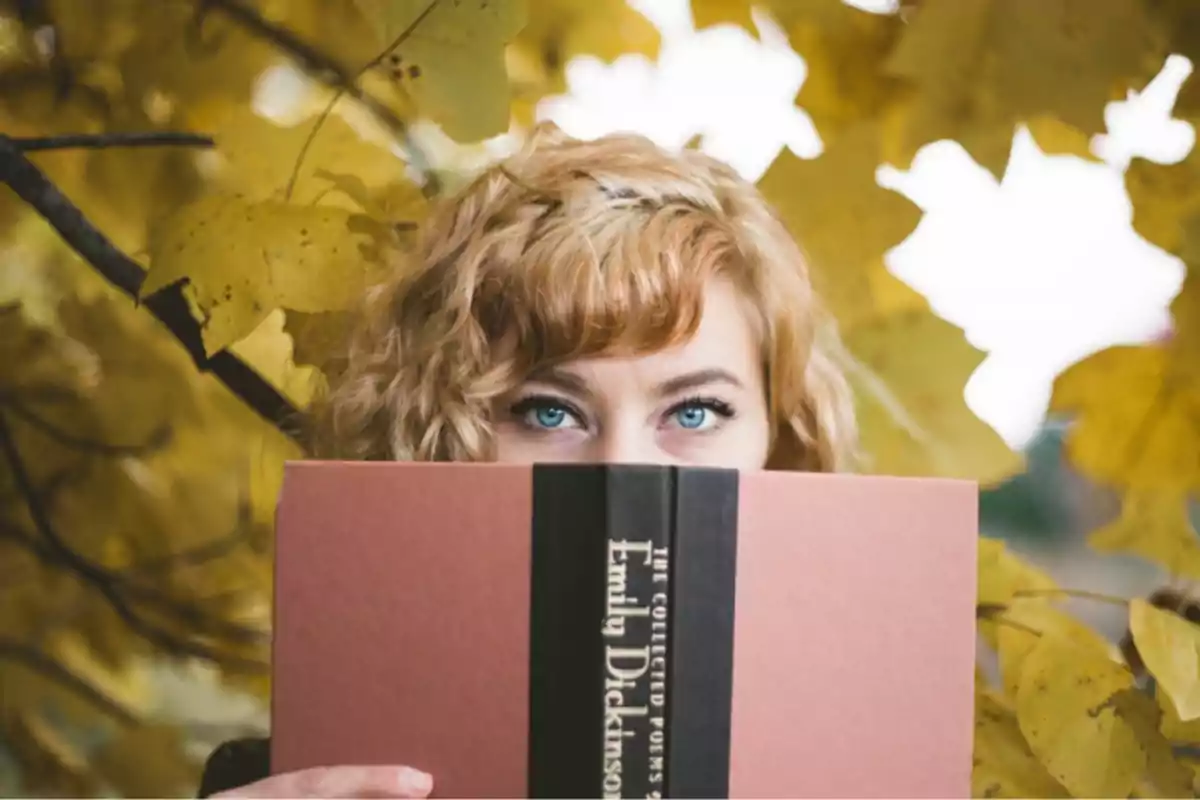 Una mujer de ojos azules y cabello rubio rizado sostiene un libro de Emily Dickinson frente a su rostro, con hojas amarillas de otoño en el fondo.