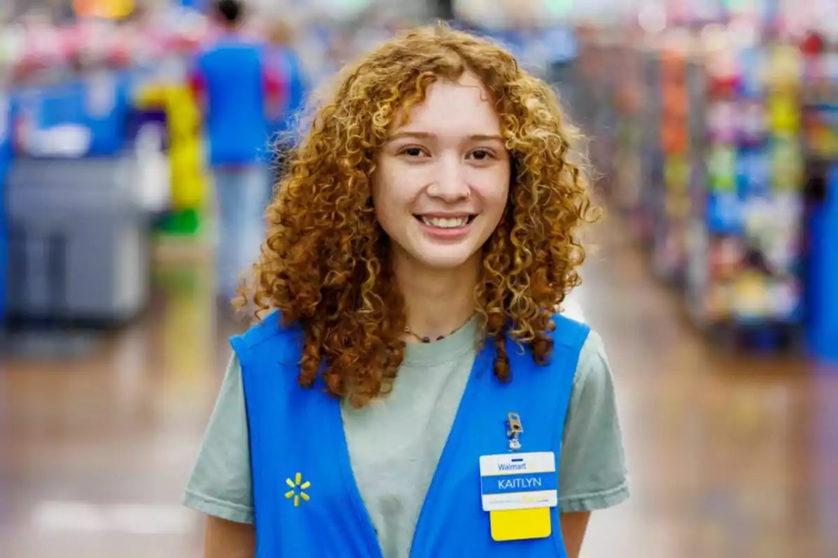 A person with curly hair and a blue uniform smiles in the aisle of a Walmart store.